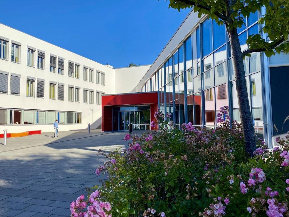 Hvit skolebygning i sollys med rød hovedinngang sentralt i bildet. Roser i bed i forgrunnen